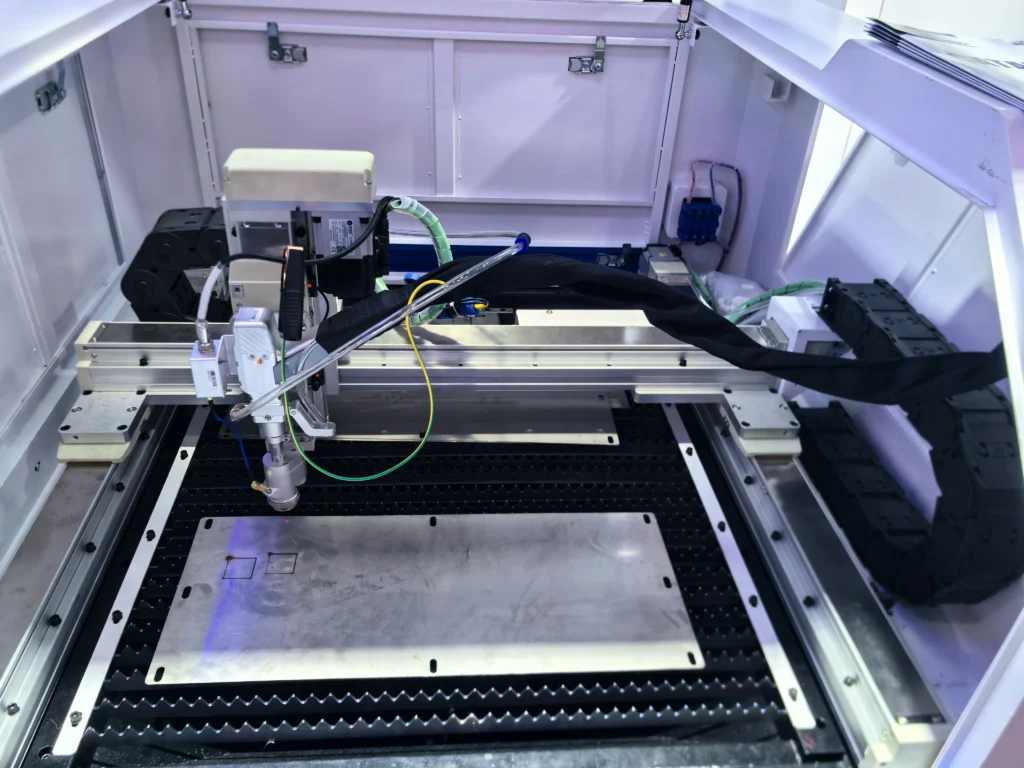 A top-down view showcasing the precise internal mechanics of the Magick CNC laser machine. The laser cutting head is operating on a stainless steel plate positioned on a honeycomb bed, with rectangular patterns already cut out. High-precision guide rails and cable drag chains are clearly visible.